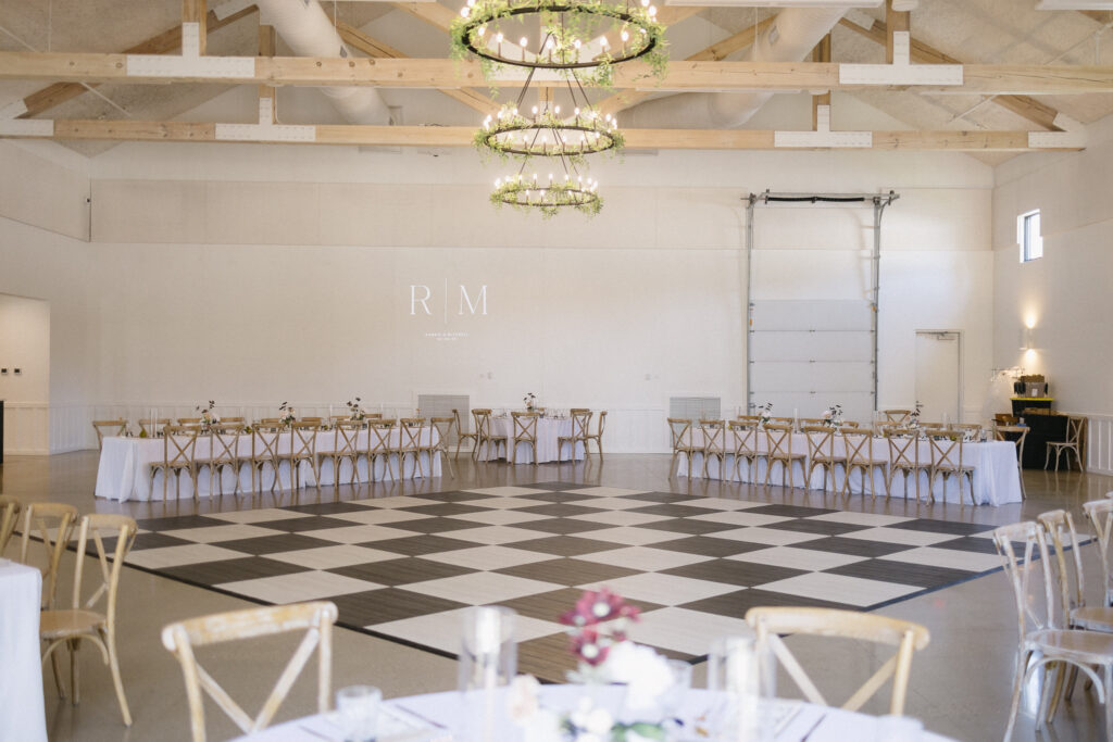 A spacious, bright event hall with high ceilings, two chandeliers, and a large checkered dance floor—ideal for those seeking elegant Michigan wedding venues. Long tables with wooden chairs line the sides, adorned with simple floral arrangements.