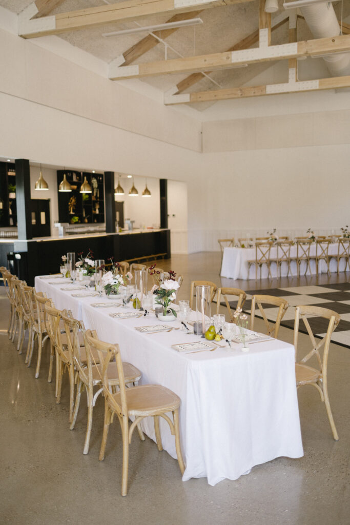 A modern event space, perfect for wedding venues, with two long tables covered in white tablecloths, set with plates, glasses, and floral centerpieces. Wooden chairs surround the tables; pendant lights and a bar are visible in the background.