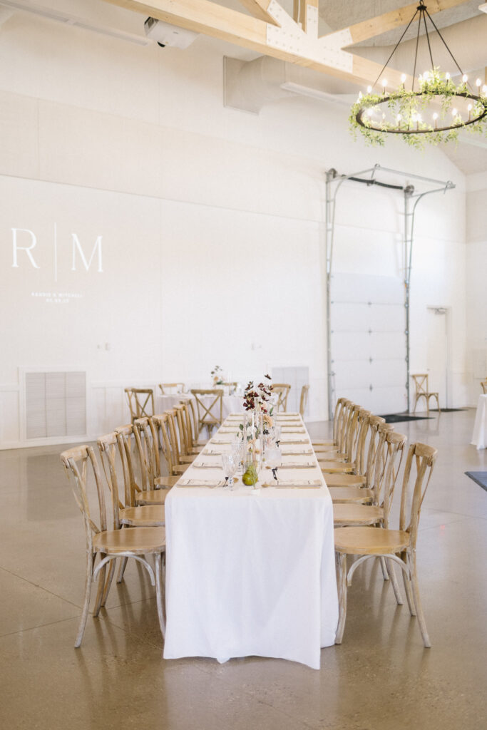 A long banquet table with a white tablecloth is set for a meal, surrounded by wooden chairs in a bright, airy room—perfect for those seeking elegant Kalamazoo wedding venues with minimalistic decor and beautiful floral arrangements.