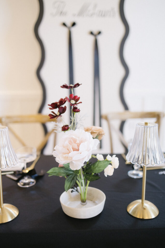 A floral centerpiece with white and dark red flowers sits on a black tablecloth, flanked by two gold lamps. Perfect for elegant wedding venues in Kalamazoo, Michigan, with wooden chairs and a white wall with black ribbon accents.