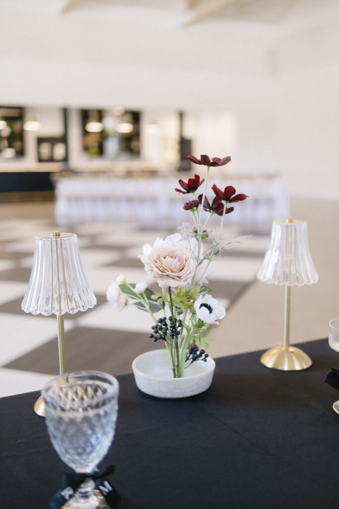A small floral arrangement with blush and burgundy flowers sits between two glass lamps on a black tablecloth, with a crystal glass nearby—an elegant setting often seen at top wedding venues in Kalamazoo, Michigan.