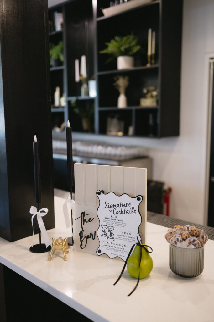 A modern bar counter with black candles, a gold dog figurine, limes, a cocktail menu, and a cup of cookies—perfect for Michigan wedding venues. Shelves with plants and glassware are visible in the background.