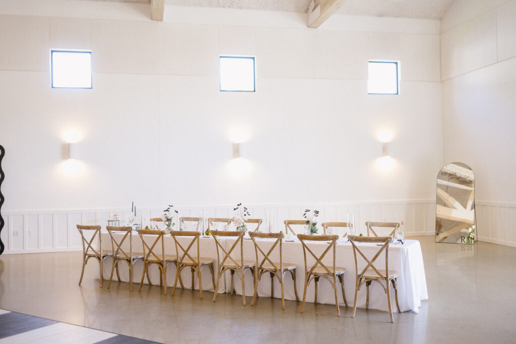 A long table with a white tablecloth and wooden chairs is set for a meal in a bright, minimalist room—an elegant choice for wedding venues or intimate gatherings, perfect for those seeking unique Kalamazoo wedding venues.