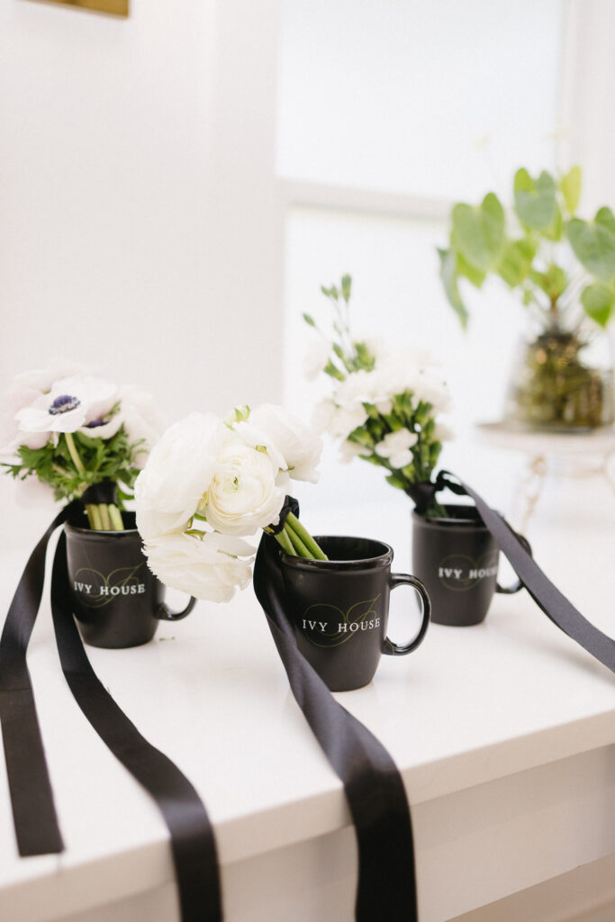 Three black mugs labeled IVY HOUSE hold white flowers and black ribbons, arranged on a white surface with a green potted plant and bright window—the perfect inspiration for elegant decor at Michigan wedding venues.