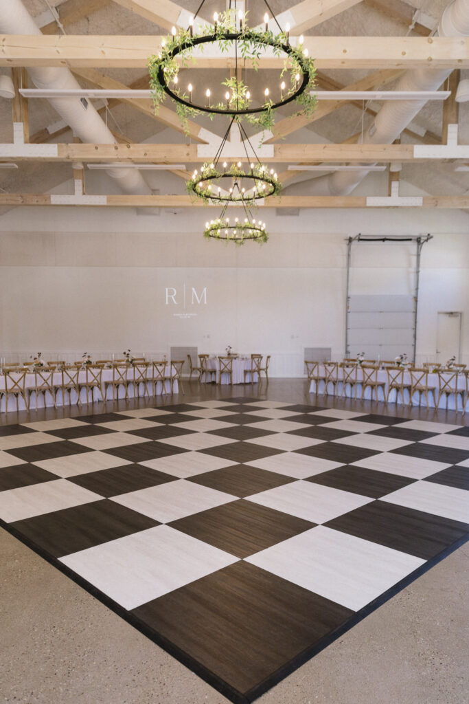 A large indoor Kalamazoo wedding venue with a black-and-white checkered dance floor, wooden ceiling beams, round chandeliers adorned with greenery, and banquet tables with chairs arranged along the back and sides of the room.
