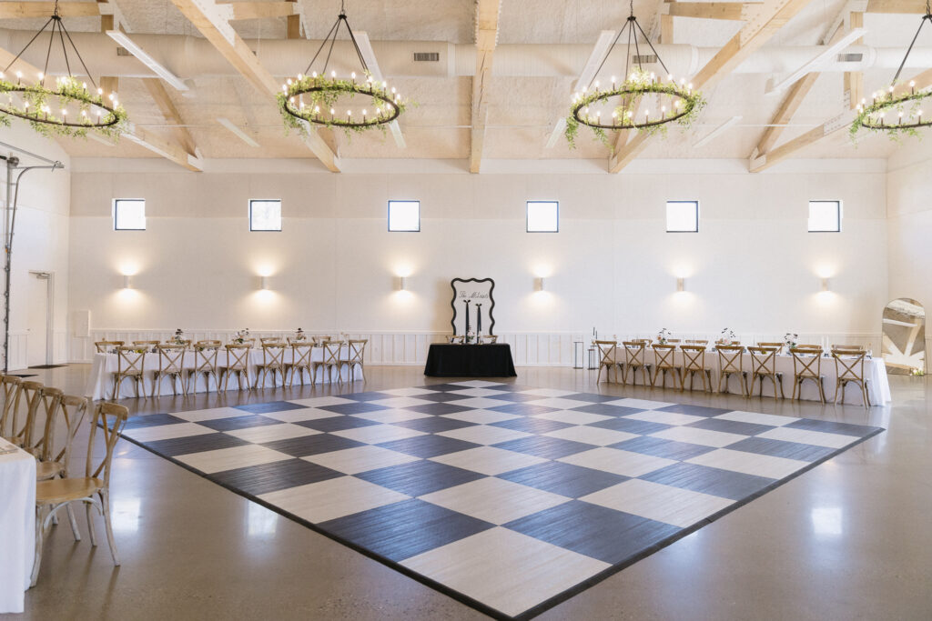 A spacious, elegant event hall—ideal among Kalamazoo wedding venues—features a large black-and-white checkered dance floor, wooden chairs and tables set along the walls, modern chandeliers with greenery, and soft natural light from small windows.