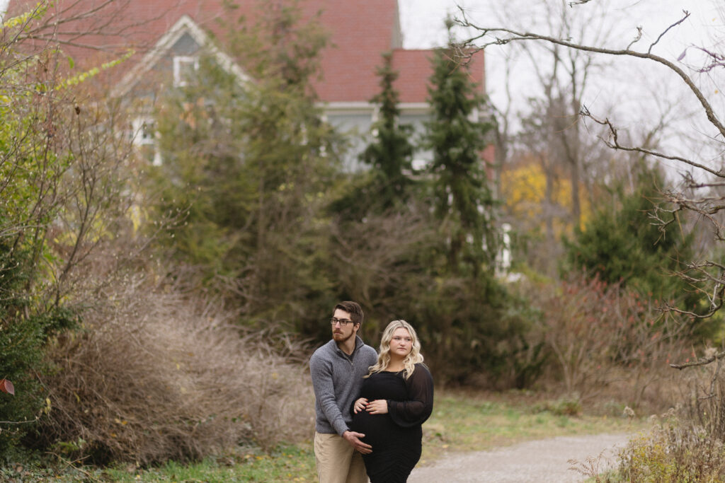 A man and a pregnant woman stand together on a garden path, surrounded by trees and shrubs, with a house and overcast sky behind them—a heartwarming scene perfect for any Michigan Family Photoshoot.