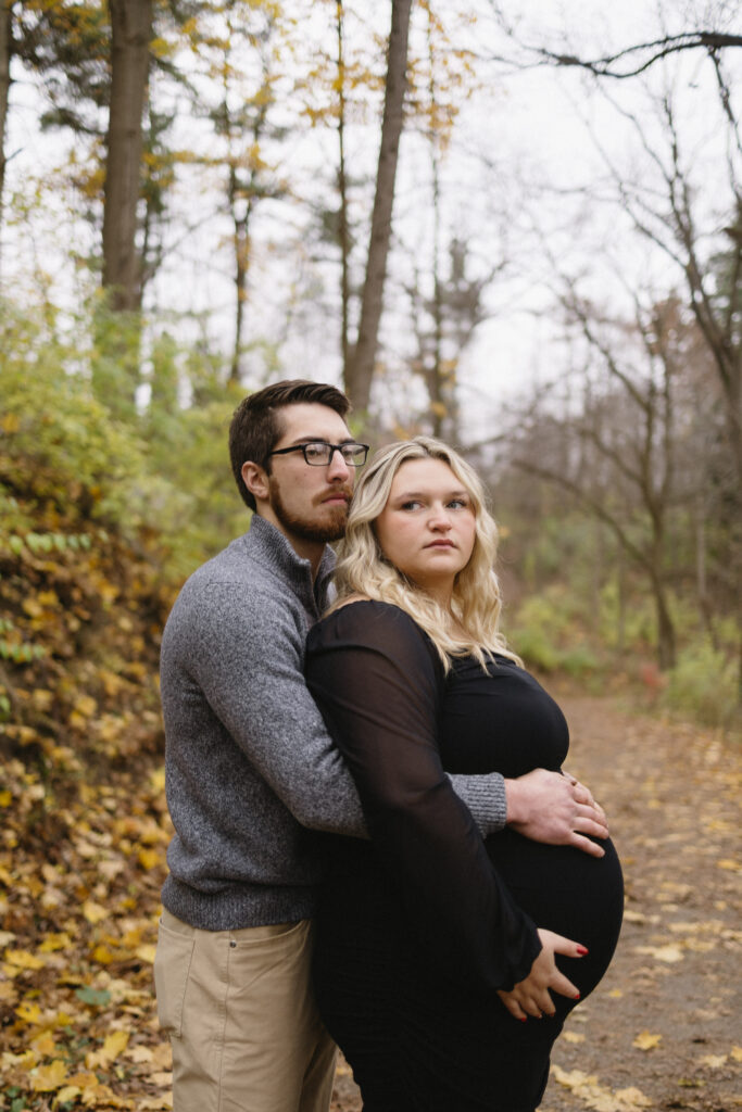 A man hugs a pregnant woman from behind on a leaf-covered path in a Michigan forest. Both have serious expressions, perfect for a heartfelt Michigan Family Photoshoot amid autumn foliage. She wears black and gently holds her belly.