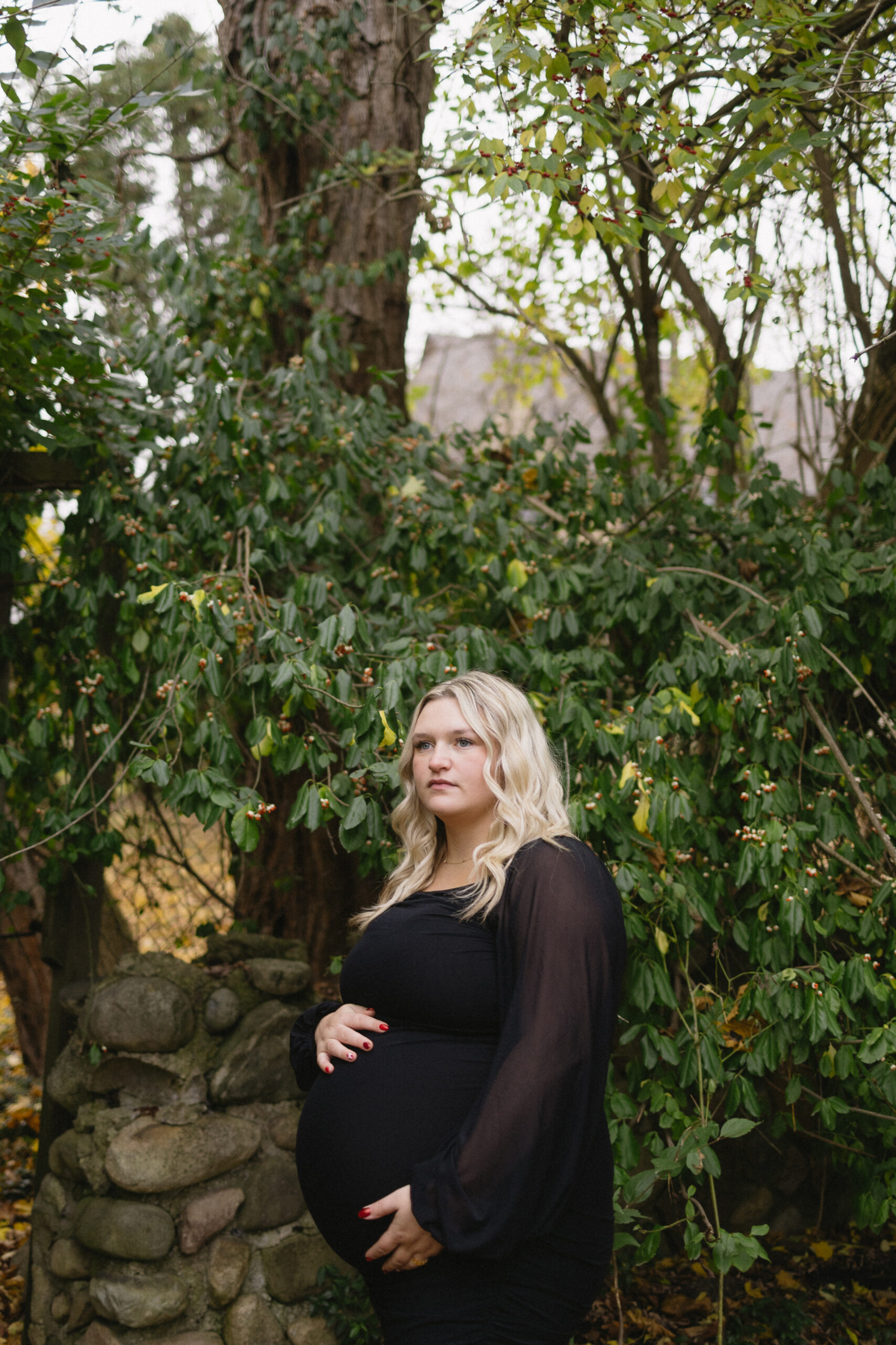 A pregnant woman with long blonde hair, wearing a black dress, stands outdoors in front of a leafy green background and stone wall, gently holding her belly and looking to the side—perfect inspiration for a Michigan Family Photoshoot.