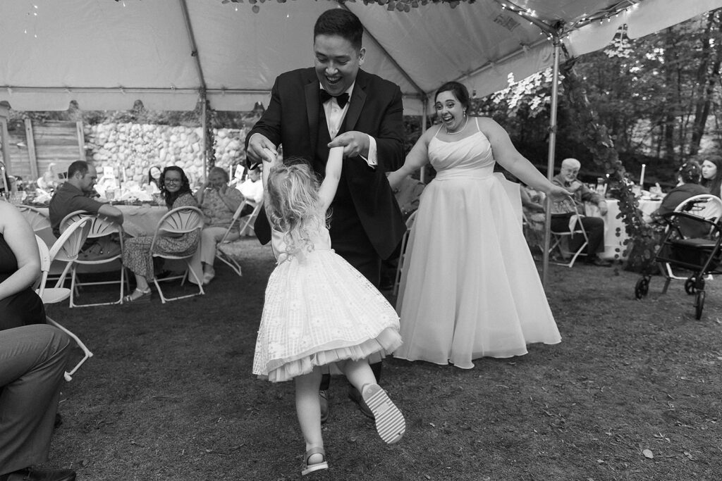 A man in a tuxedo spins a young girl in a white dress while a woman in a gown smiles nearby. They are under a tent at an outdoor event, with guests seated at decorated tables in the background.