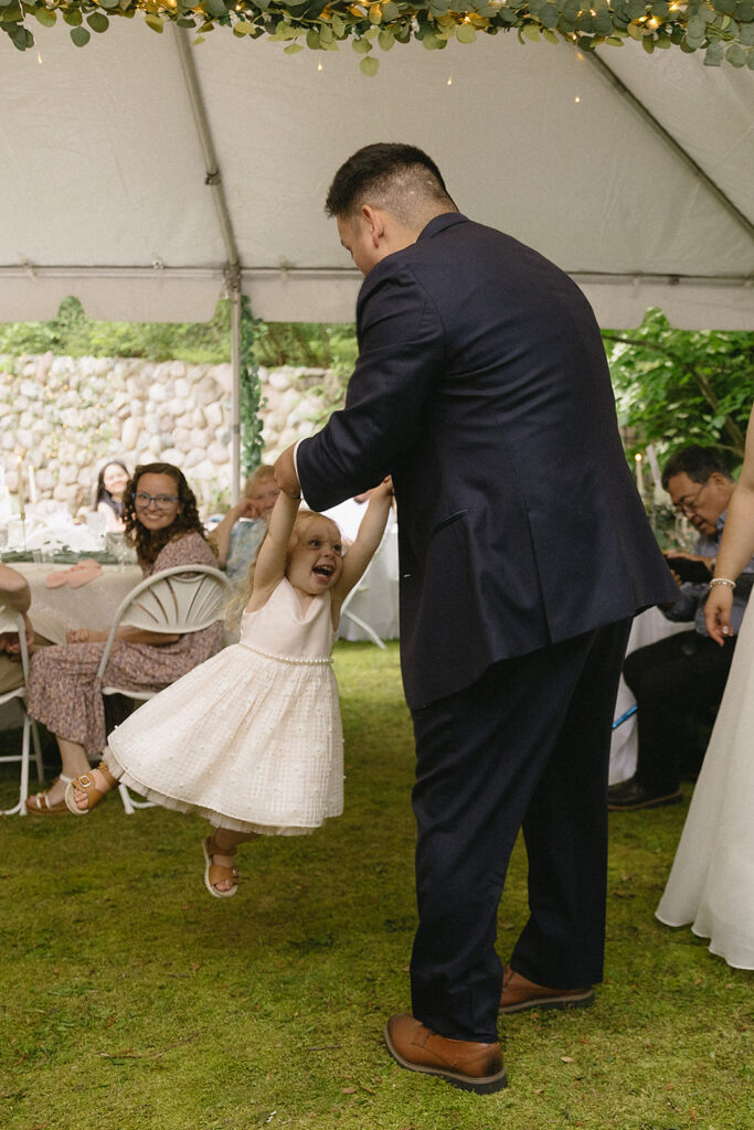 A man in a navy suit swings a smiling young girl in a white dress by her arms under a tent at an outdoor event, while seated guests watch and smile in the background.
