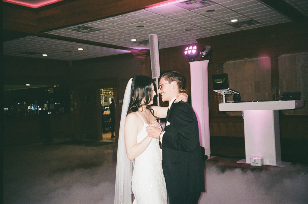 A bride and groom share a first dance on a fog-covered dance floor in a dimly lit reception hall, gazing into each other’s eyes and smiling. The bride wears a white dress and veil, and the groom is in a black tuxedo.