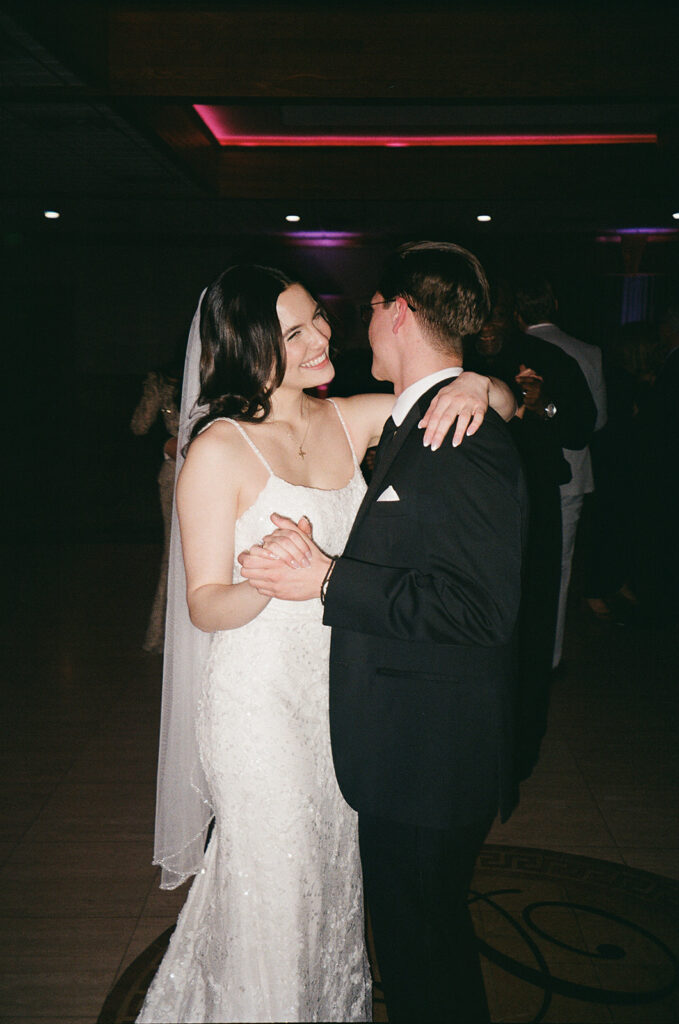 A bride in a white dress and veil smiles as she dances with a groom in a black suit and glasses on a dimly lit dance floor, likely during their wedding reception.