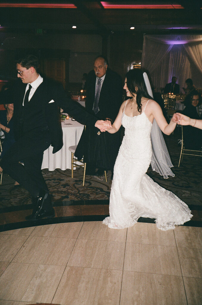 A bride in a white gown and veil and a groom in a black suit and tie hold hands while dancing at their wedding reception, surrounded by guests seated at decorated tables.
