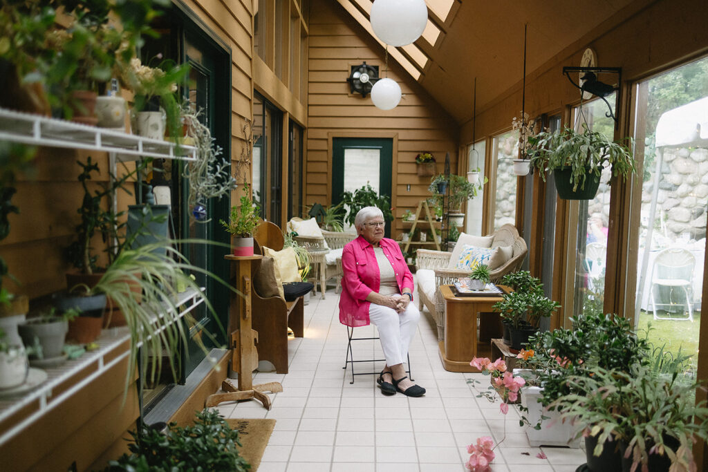 An elderly woman with white hair, wearing a pink jacket and white pants, sits on a chair in a bright sunroom filled with potted plants, cozy furniture, and large windows looking out onto a green garden.