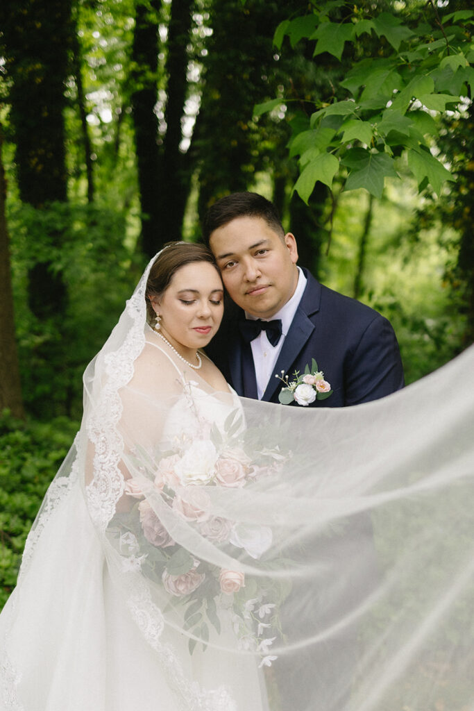 A bride and groom pose together in a lush, green outdoor setting. The bride holds a bouquet of pink and white flowers, her lace veil draped around them both, while the groom embraces her in a navy suit and bow tie.
