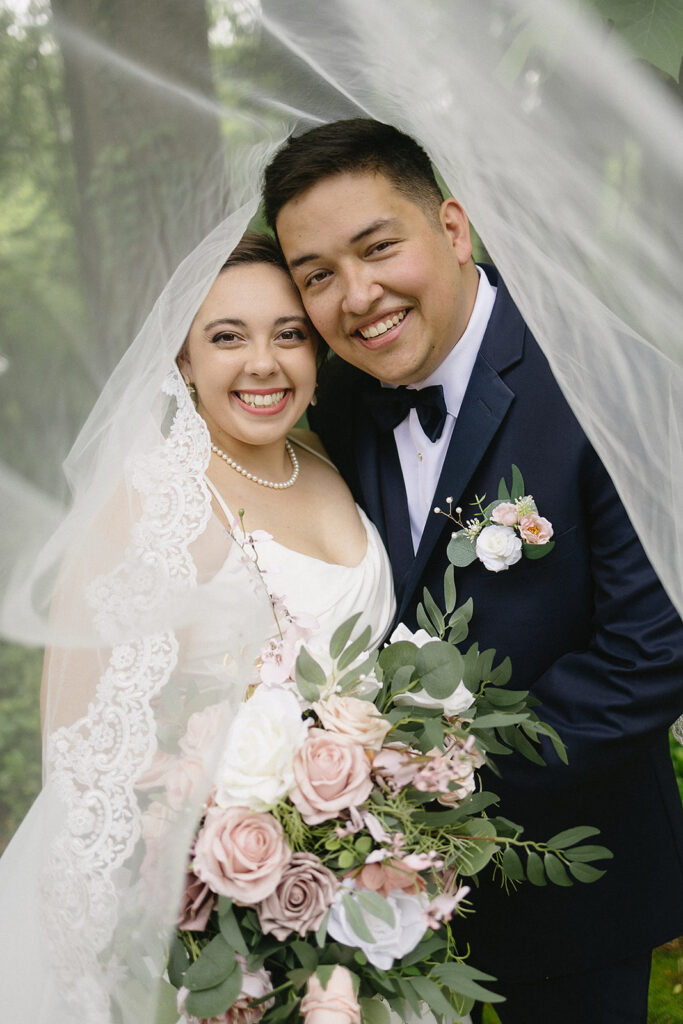 A smiling bride and groom pose closely together outdoors, framed by the bride’s veil. The bride holds a bouquet of pink and white flowers with greenery, and both wear formal wedding attire.