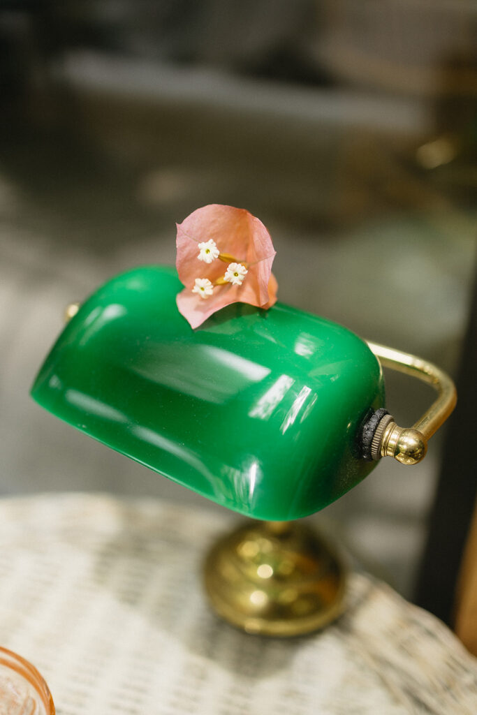 A green glass desk lamp with a brass base sits on a wicker surface, topped with a small pink flower and three tiny white blossoms.