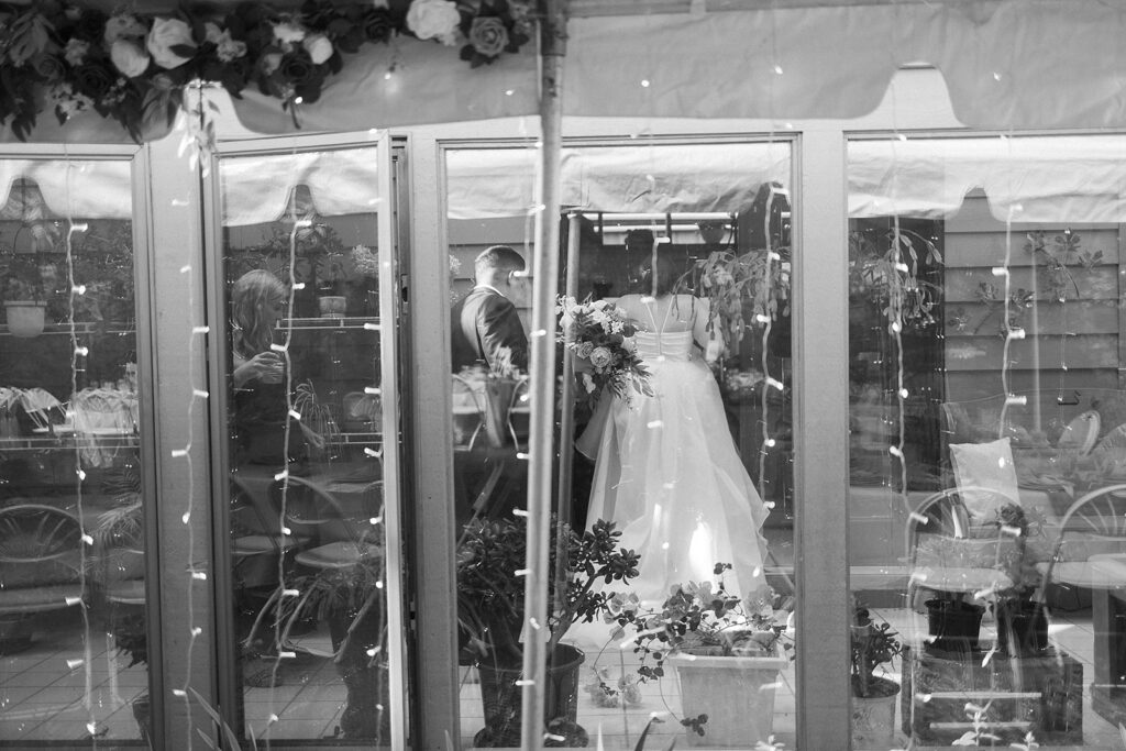 A bride and groom stand together indoors, seen through glass doors decorated with string lights and flowers. The scene is framed by plants and reflections, creating an intimate, romantic atmosphere.