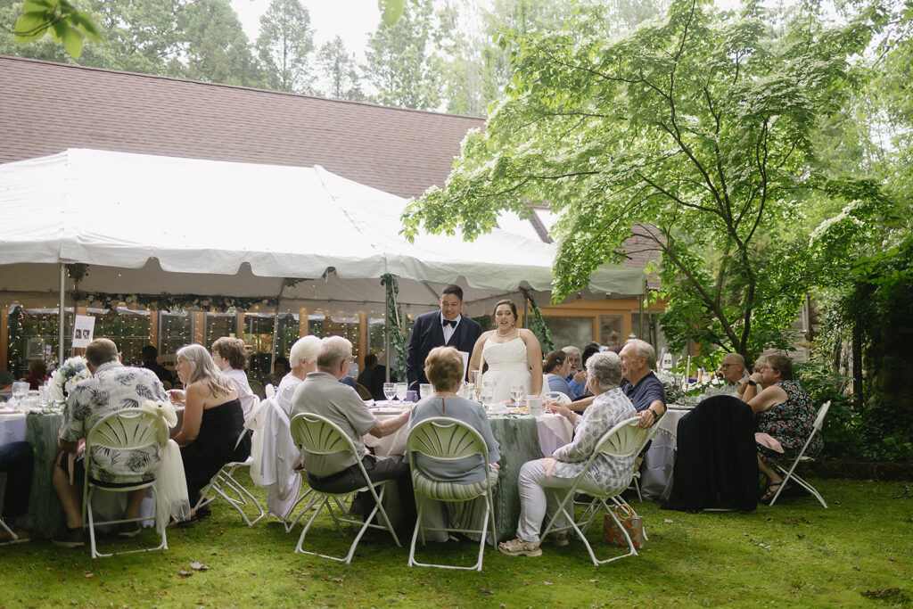 A bride and groom stand beside a table, addressing seated guests at an outdoor wedding reception under a white tent surrounded by greenery.