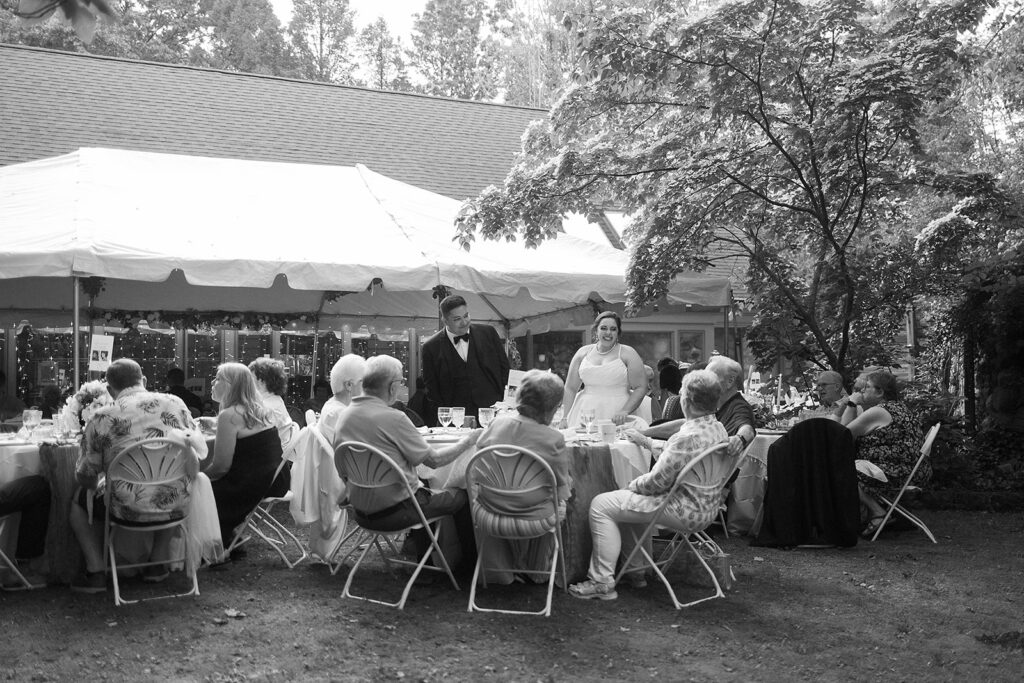 A bride and groom stand smiling near a table, surrounded by seated guests at an outdoor wedding reception under a canopy, with trees and string lights in the background.