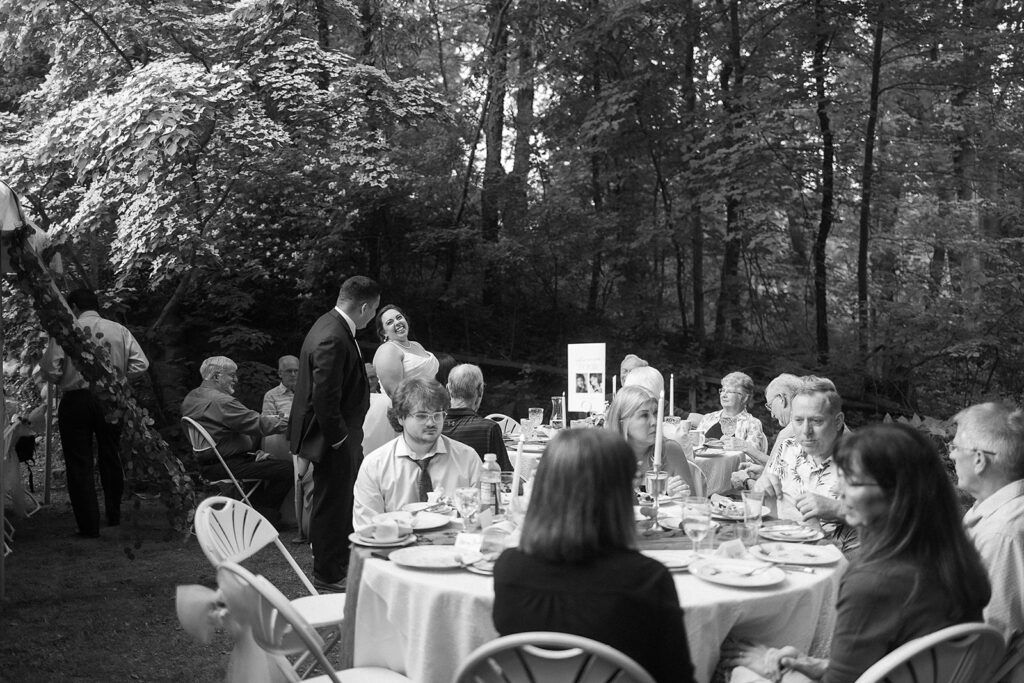 A black-and-white photo of people gathered outdoors at round tables, sharing a meal under trees. A couple stands and smiles in the background, while guests sit and converse at a table in the foreground.