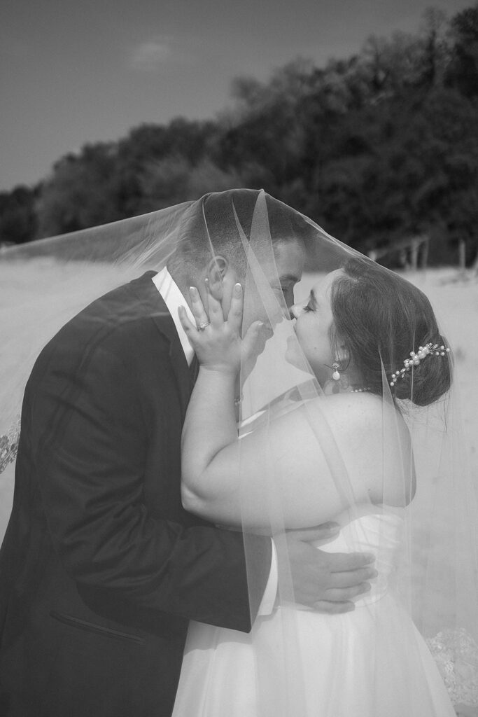 A bride and groom stand close together outdoors, embracing and touching foreheads under a sheer wedding veil, with trees in the background. The image is in black and white.