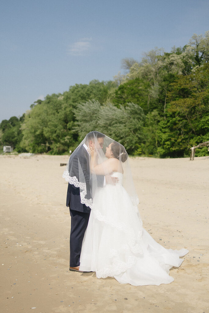 A bride and groom stand close together on a sandy beach, facing each other and smiling under a lace-edged wedding veil, with green trees and blue sky in the background.