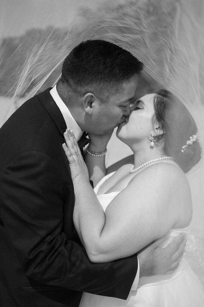 A bride and groom share a kiss under a translucent veil. The bride wears a pearl necklace and earrings, and the groom is in a dark suit. The intimate moment is captured in black and white.