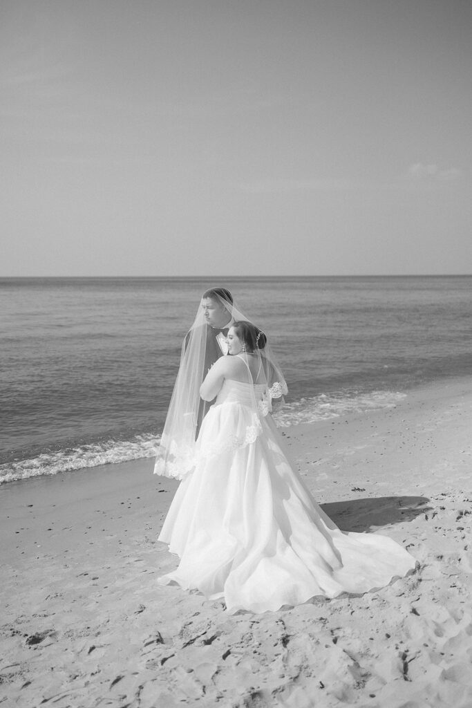 A bride in a wedding dress and veil stands on a sandy beach, facing the ocean, with gentle waves and a clear sky in the background. The scene is captured in black and white.