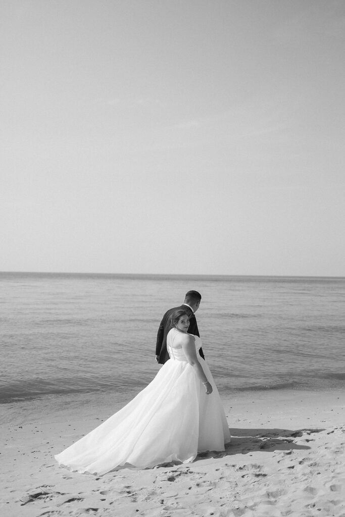 A bride in a flowing white gown stands on a sandy beach, looking back over her shoulder, while a groom in a suit stands beside her, both facing the calm sea under a clear sky. The image is in black and white.