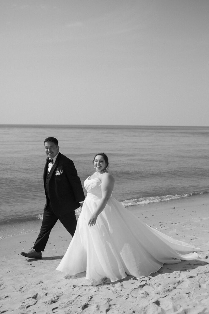 A bride in a flowing gown and a groom in a suit walk hand in hand along a sandy beach, smiling, with calm ocean water in the background. Black and white photo.
