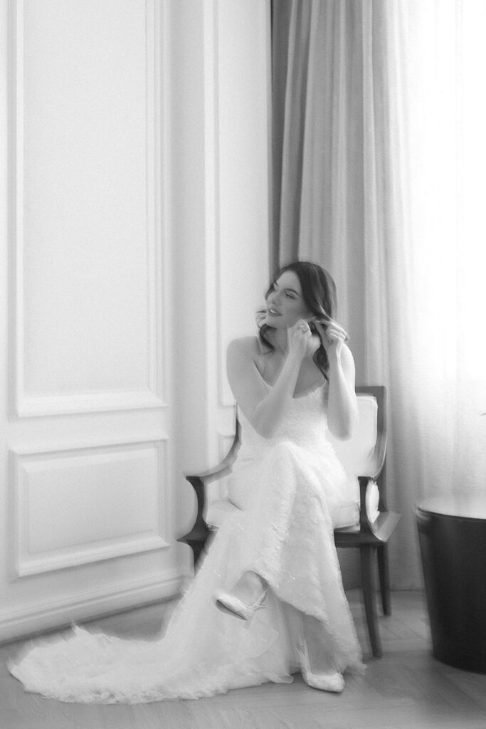 A bride in a white wedding dress sits on a chair by a window, putting on earrings. The image is softly focused, creating an ethereal and elegant atmosphere.