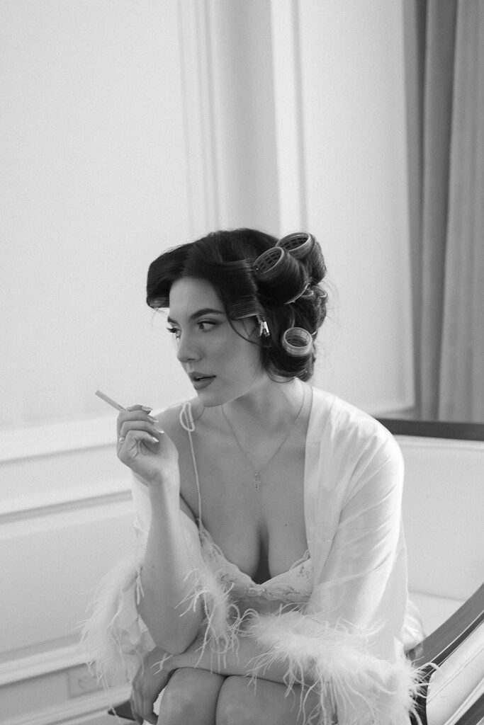 A woman with hair rollers, wearing a light robe with feathered sleeves, sits on a chair holding a cigarette. She looks to the side thoughtfully in a softly lit, elegant room. The photo is in black and white.