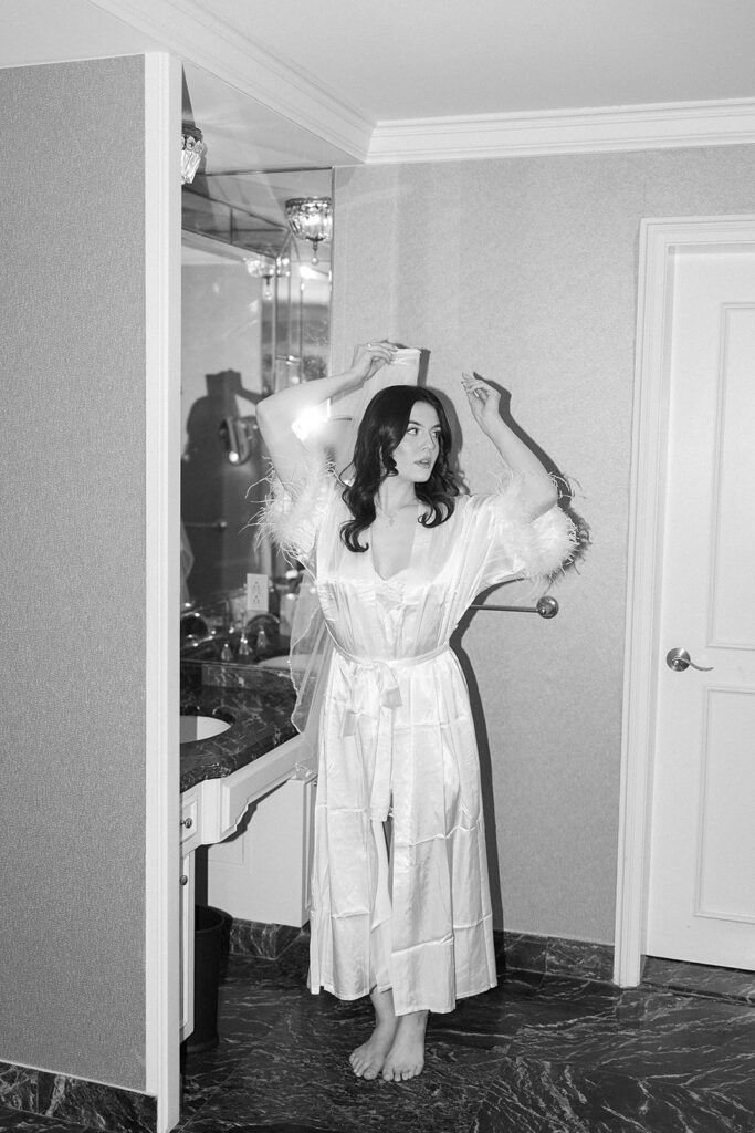 A woman in a white satin robe with feathered sleeves stands barefoot in a marble-floored bathroom, facing slightly to the side with her arms raised, next to a vanity mirror. The image is in black and white.