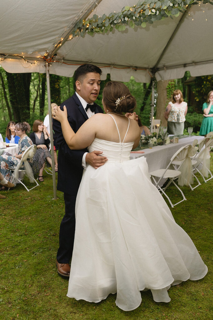 A bride and groom share their first dance under a white tent at an outdoor wedding. The bride wears a white gown; the groom is in a dark suit. Guests seated at tables watch in the background, surrounded by greenery.