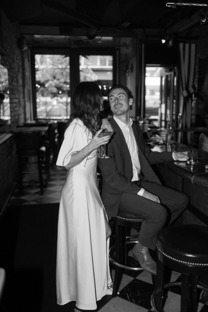 A woman in a white dress stands beside a man in a suit seated at a bar, holding a wine glass and leaning in closely as they share a joyful, intimate moment in a dimly lit restaurant.