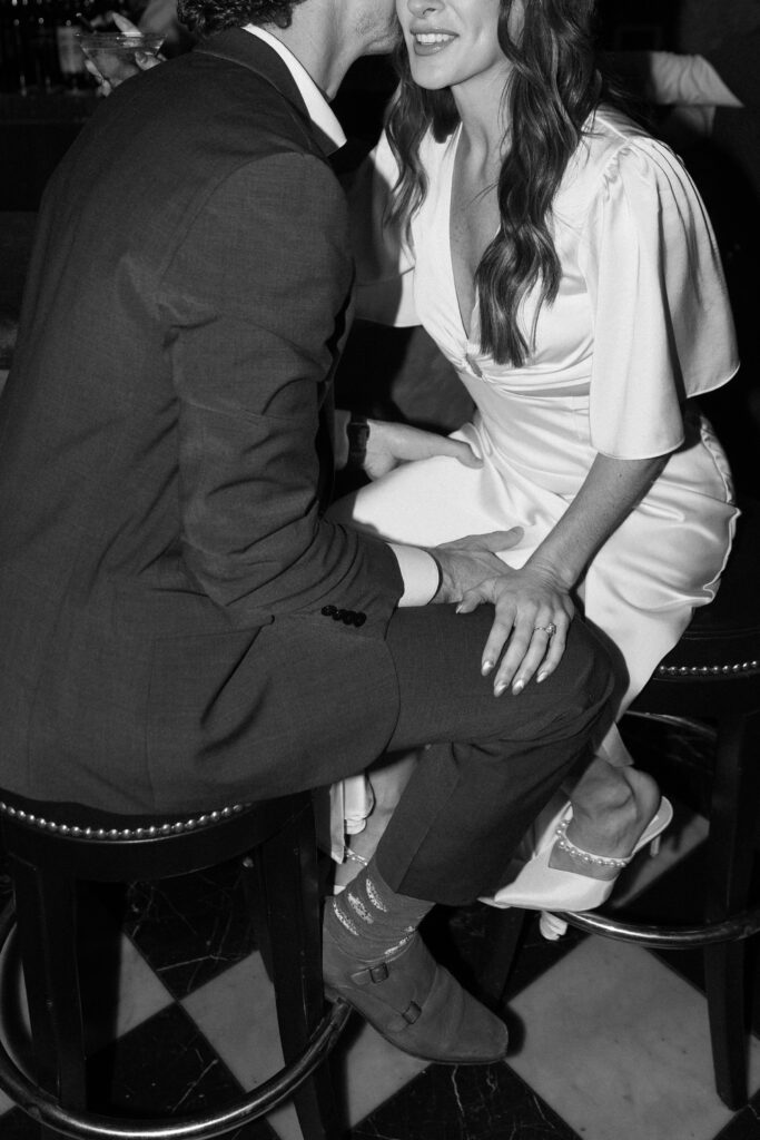 A man in a suit whispers to a smiling woman in a white dress as they sit closely on bar stools, their hands touching. The image is in black and white.
