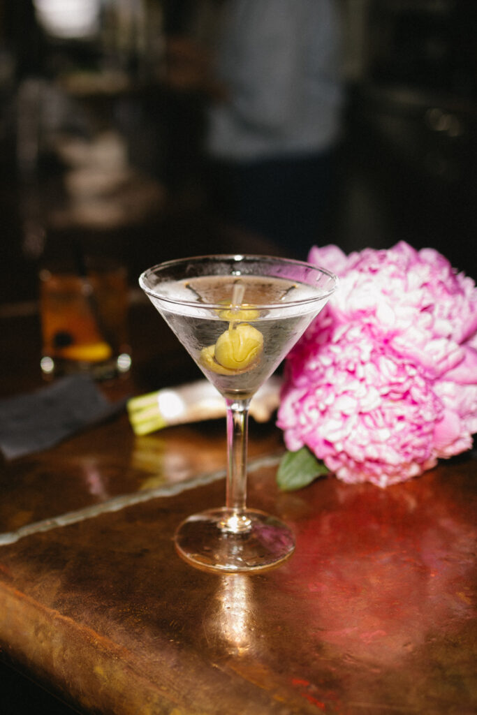 A martini with an olive garnish sits on a wooden bar next to a pink peony flower, with a dark cocktail and a napkin in the blurred background.