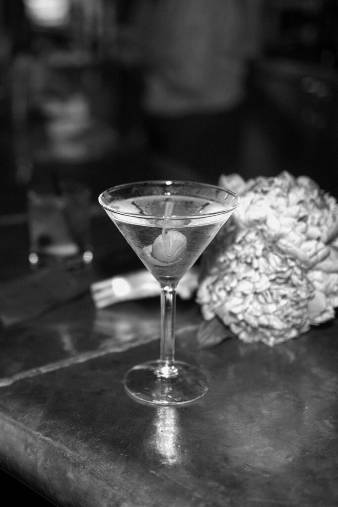 A martini with an olive sits on a bar next to a bouquet of flowers. The image is in black and white, with a blurred background and reflective surface.