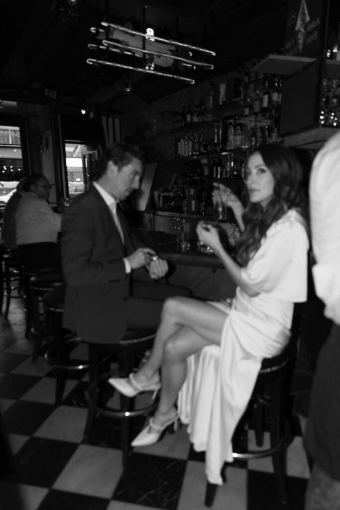 A black and white photo of a man in a suit and a woman in a white dress sitting at a bar. The woman is facing the camera, crossing her legs, while the man looks down at something in his hands. The bar scene is lively.
