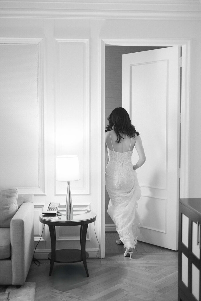 A woman in a long, elegant dress walks through an open door in a bright, stylish room with a lamp on a side table and a sofa nearby. The photo is in black and white.