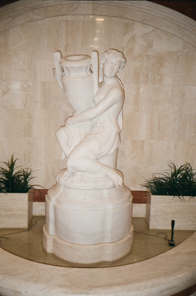 A white marble statue of a woman kneeling and holding a large amphora, set in a small indoor fountain with green plants on either side and beige stone walls in the background.
