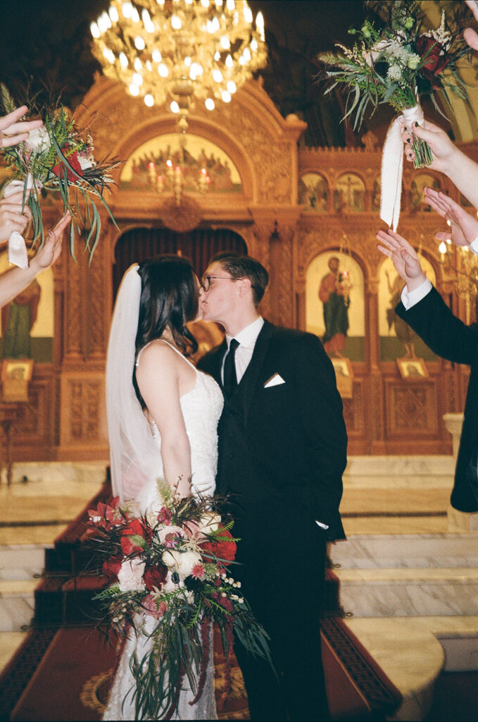 A bride and groom kiss at the altar of a church, holding bouquets of red and white flowers, while friends raise bouquets around them under a large chandelier.