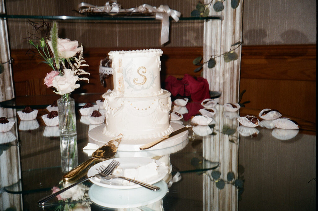 A white, two-tiered cake with a gold S sits on a glass table, partially sliced, next to a fork, cake server, and vase of flowers. Cupcakes and greenery are arranged decoratively in the background.