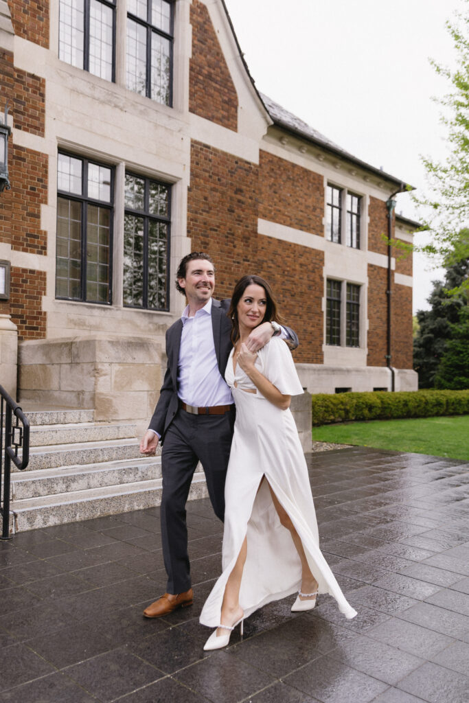 A couple dressed formally, with the woman in a white dress and the man in a suit, walk arm in arm and smiling outside a large brick building with tall windows and stone steps on a rainy day.