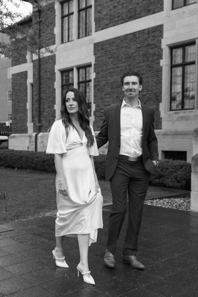 A woman in a white dress and heels walks beside a man in a suit on a paved path in front of a brick building, both looking ahead. The image is in black and white.