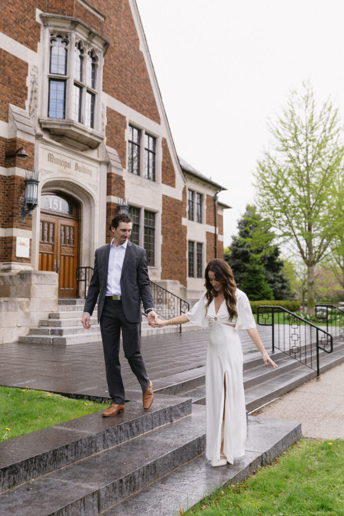 A man in a dark suit holds hands with a woman in a white dress as she carefully walks down stone steps outside a brick municipal building with arched windows on a cloudy day.