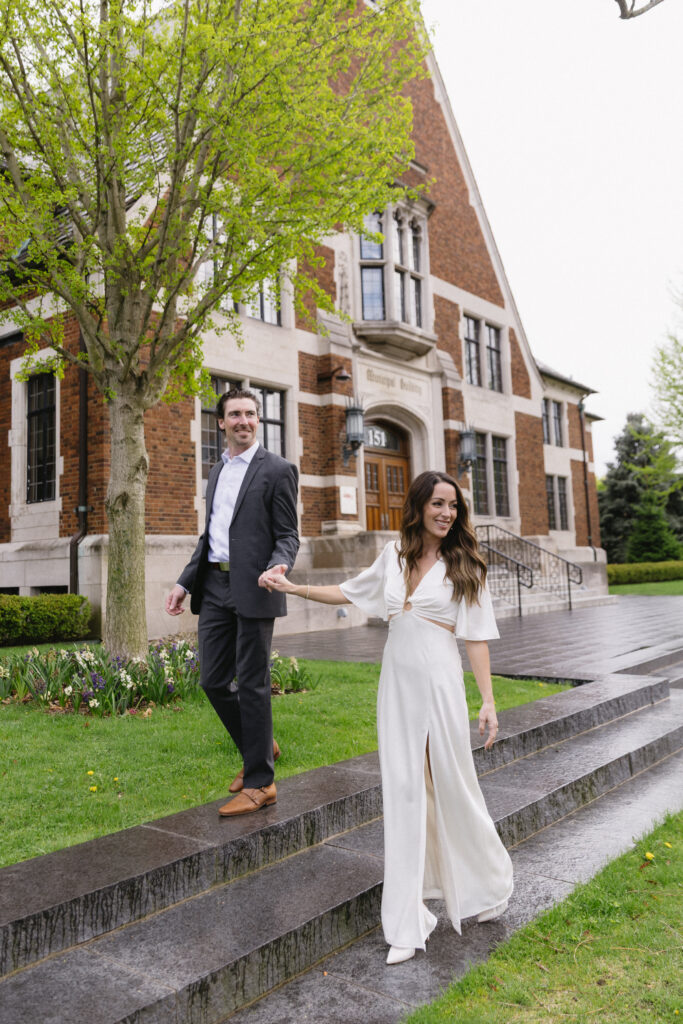 A couple dressed in formal attire, with the woman in a white dress and the man in a dark suit, walk hand in hand down wet stone steps outside a brick building surrounded by greenery.