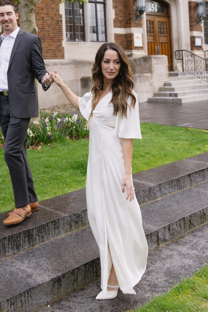 A woman in a white dress and heels walks down stone steps outside a brick building, holding hands with a man in a gray suit. She looks at the camera and smiles slightly. Flowers and greenery are visible in the background.